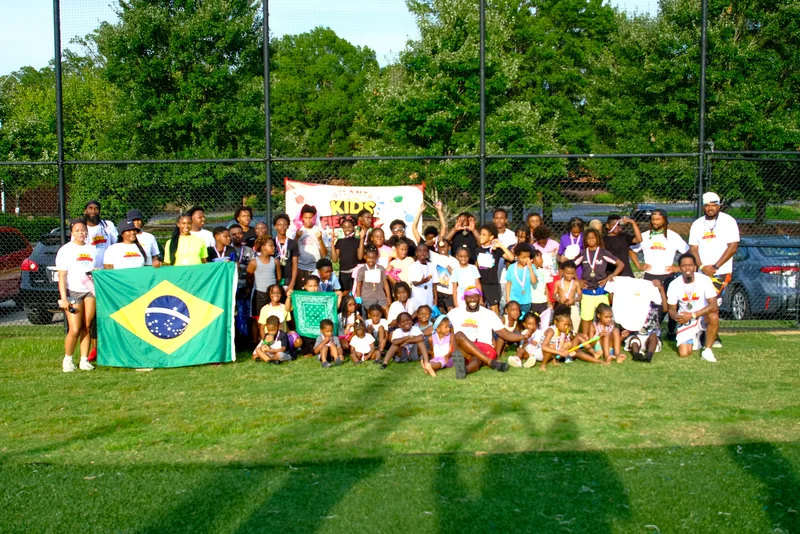 A large group of kids and coaches gather behind the Kids field day banner, with a Brazil flag held up, at a Faith First Fitness event