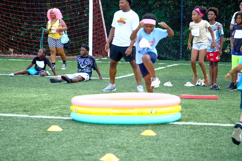A child in a headband leaps over an inflatable obstacle as kids and a coach cheer